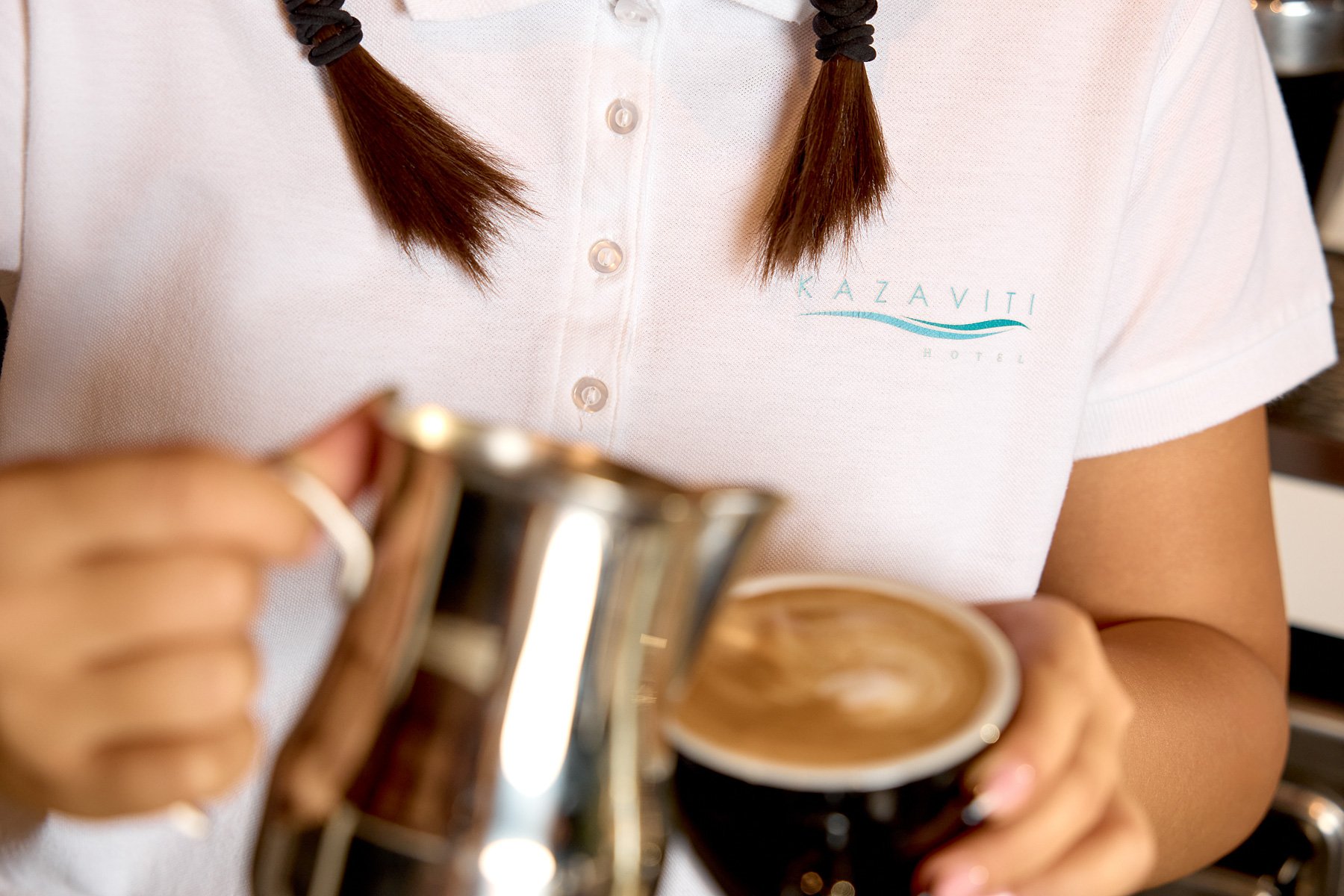 Person wearing a white Kazaviti Hotel polo shirt, pouring steamed milk from a stainless steel pitcher into a black coffee cup.