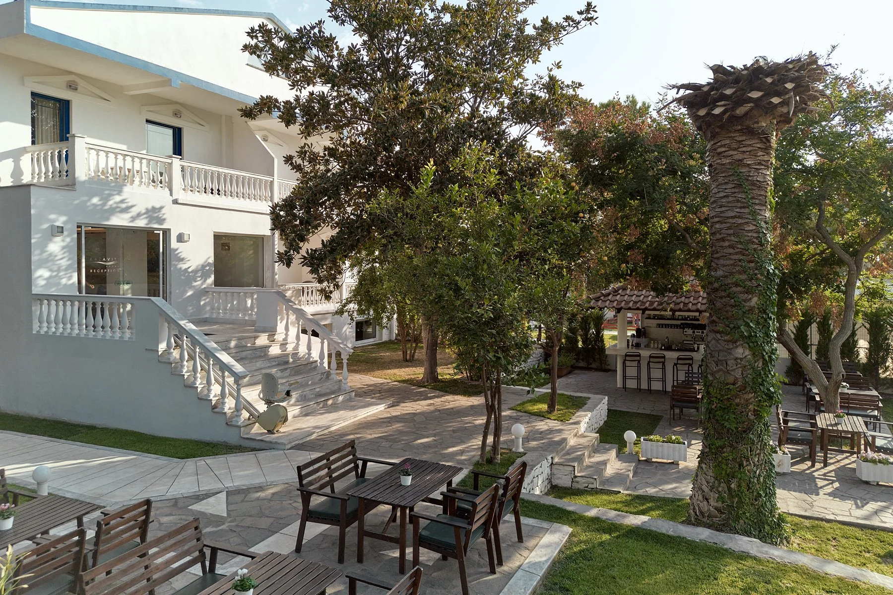 Outdoor patio area with wooden tables and chairs, surrounded by trees and a white two-story building with balconies, stairs, and a bar area with stools.