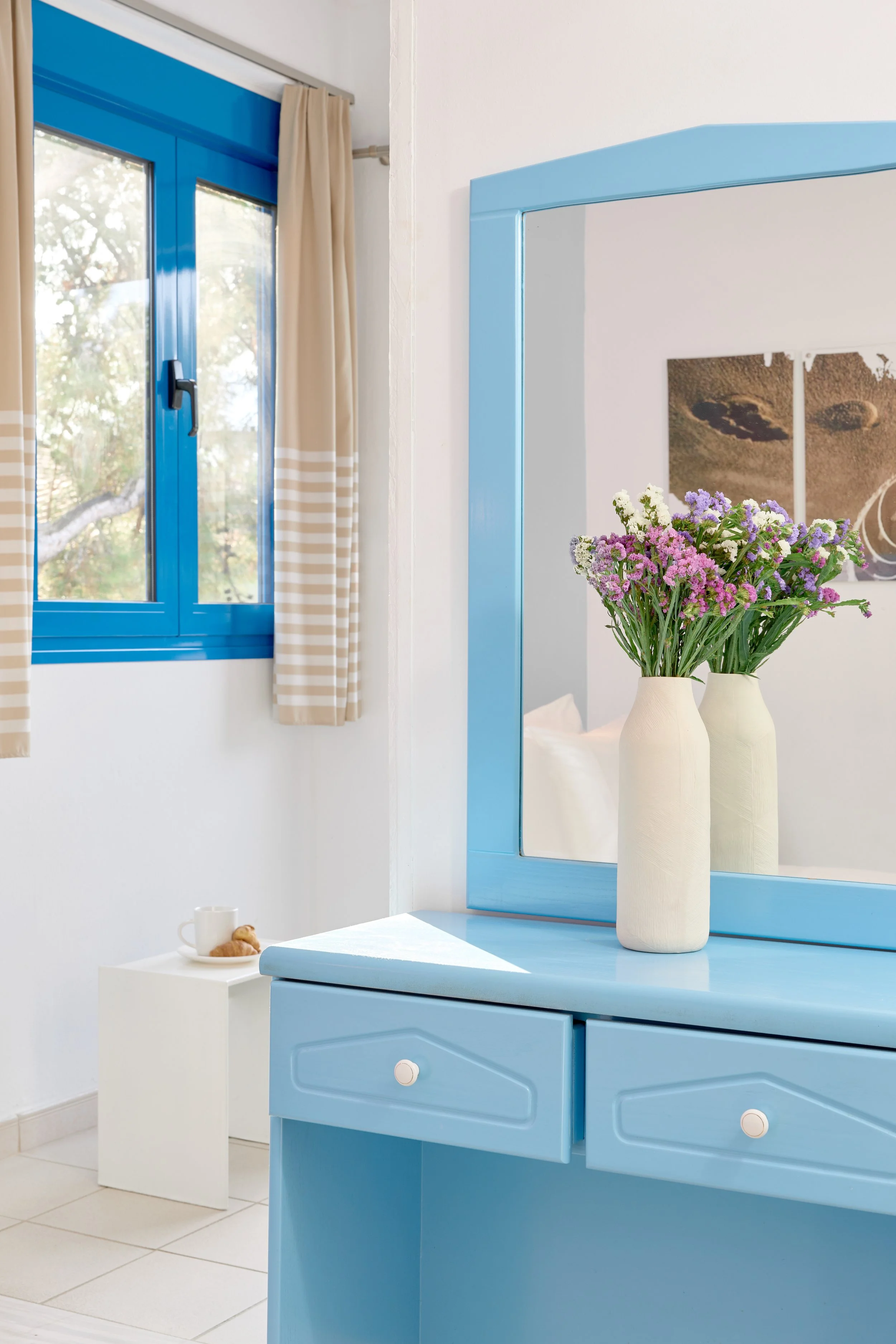A blue dresser with a mirror and a door with a window, beige and white striped curtains, and a vase with purple and white flowers on top.