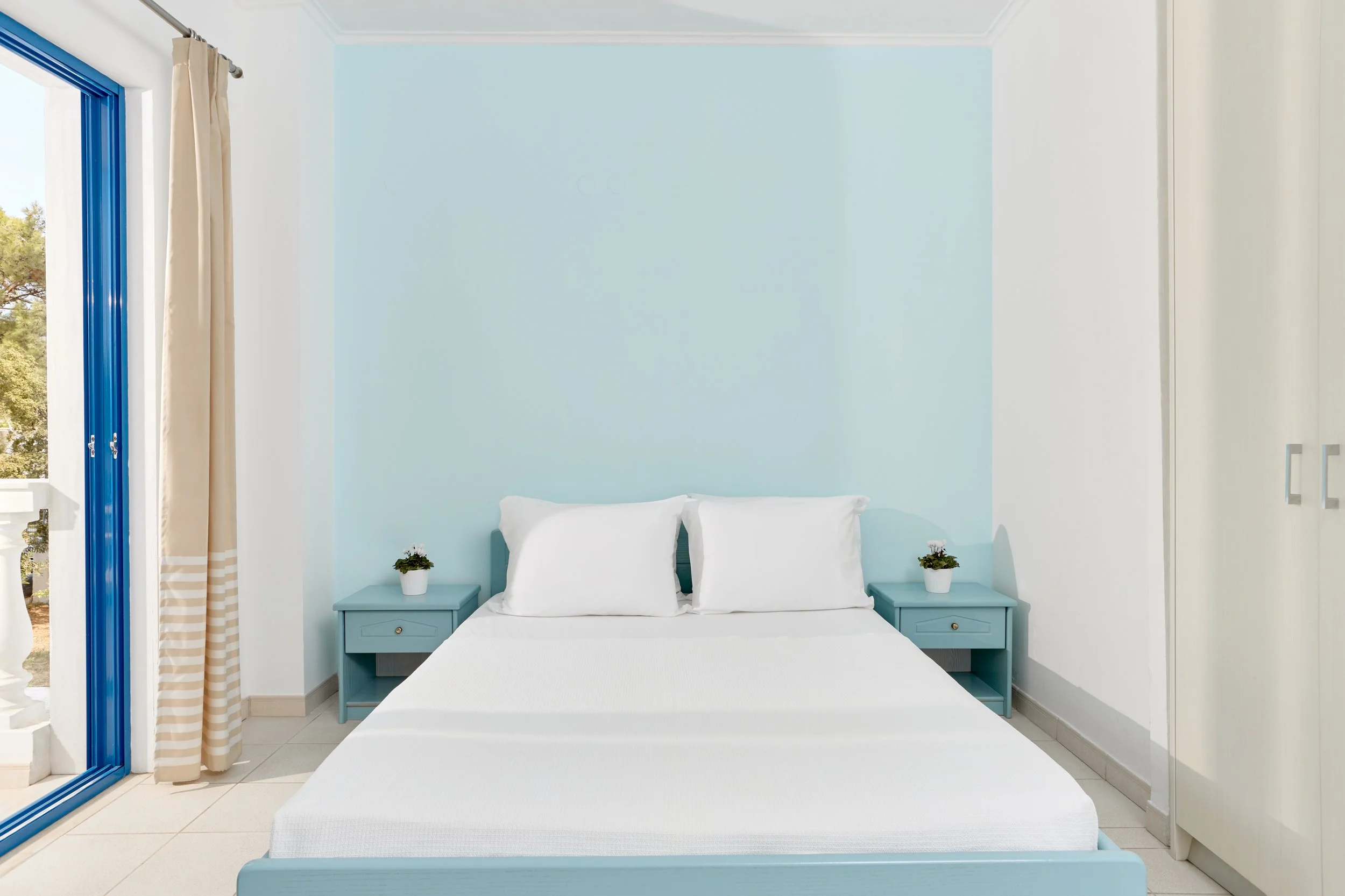 A minimalist bedroom with a white bed, two blue nightstands with small potted plants, a blue accent wall behind the bed, beige curtains, and a large sliding glass door leading outside.