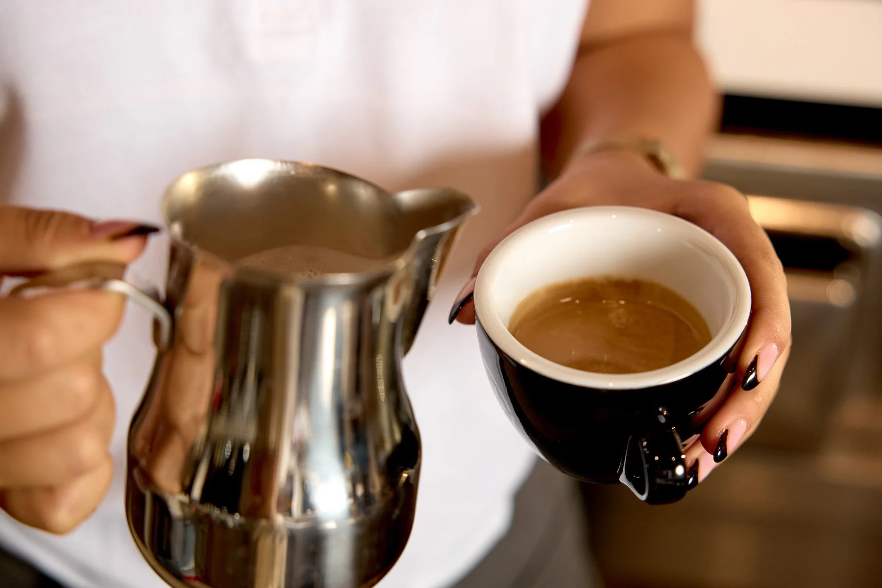 Person holding a black coffee cup in one hand and a metal creamer pitcher in the other.