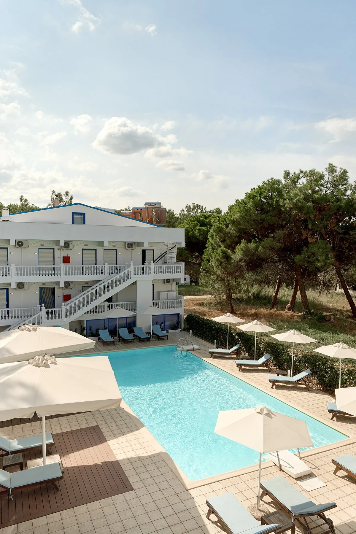 A hotel pool area with lounge chairs and umbrellas, adjacent to a white three-story building surrounded by trees.