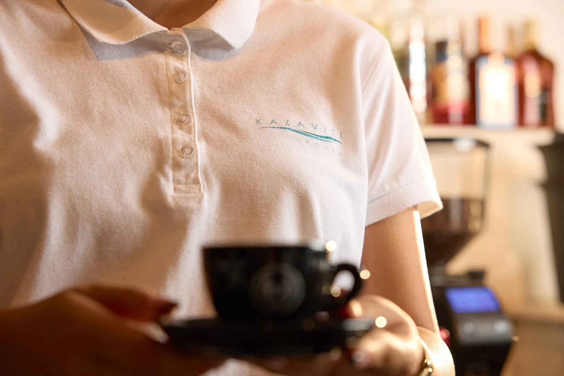A person wearing a white polo shirt with an embroidered logo 'KAZAVITI' is holding a black coffee cup in a café setting, with shelves of condiments in the background.