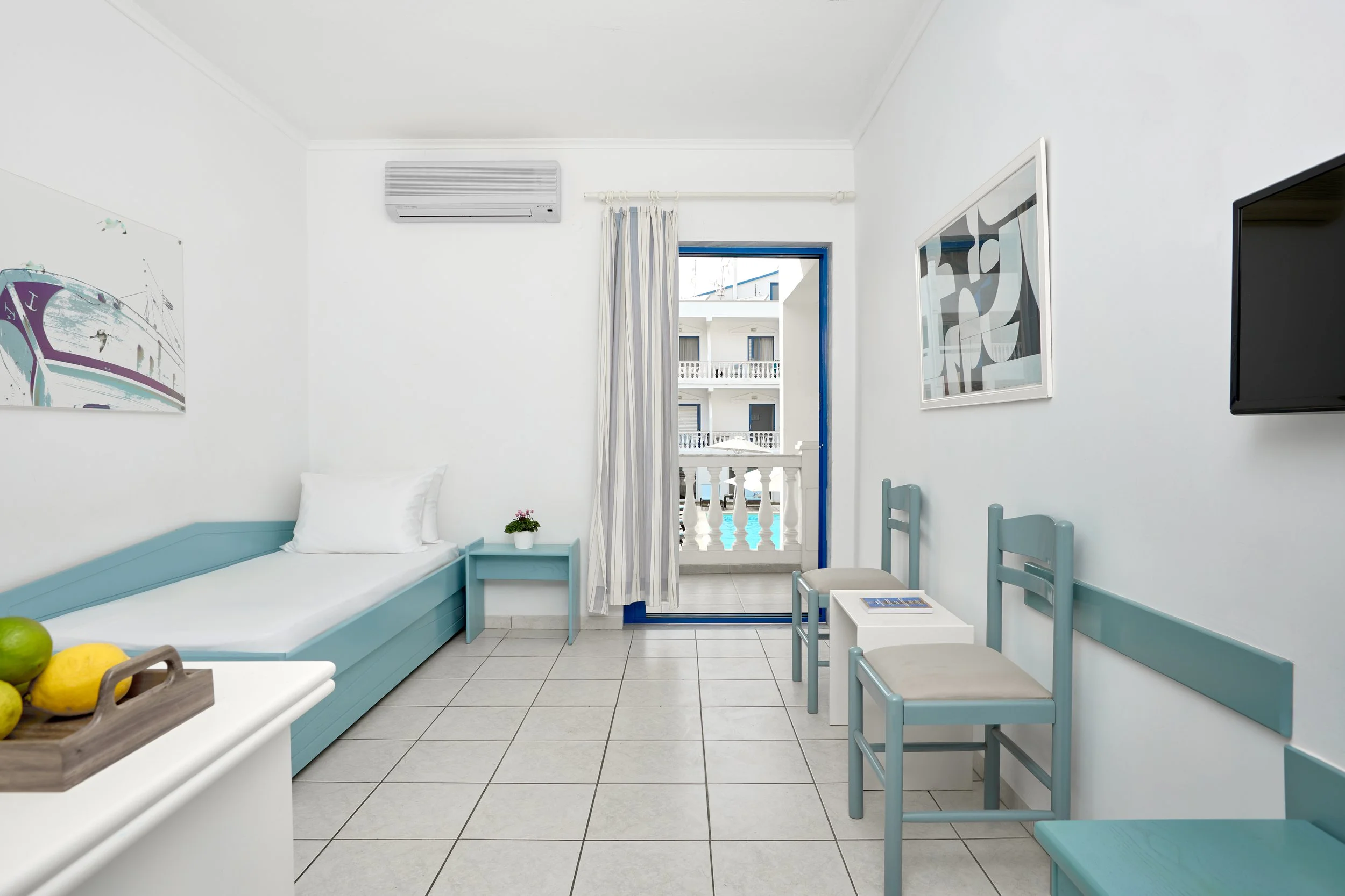 A small, bright hotel room with a white and light blue color scheme. It has a single bed, a small side table with a potted plant, a table with two chairs, a wall-mounted TV, a window with a balcony overlooking a building with balconies, and artwork o