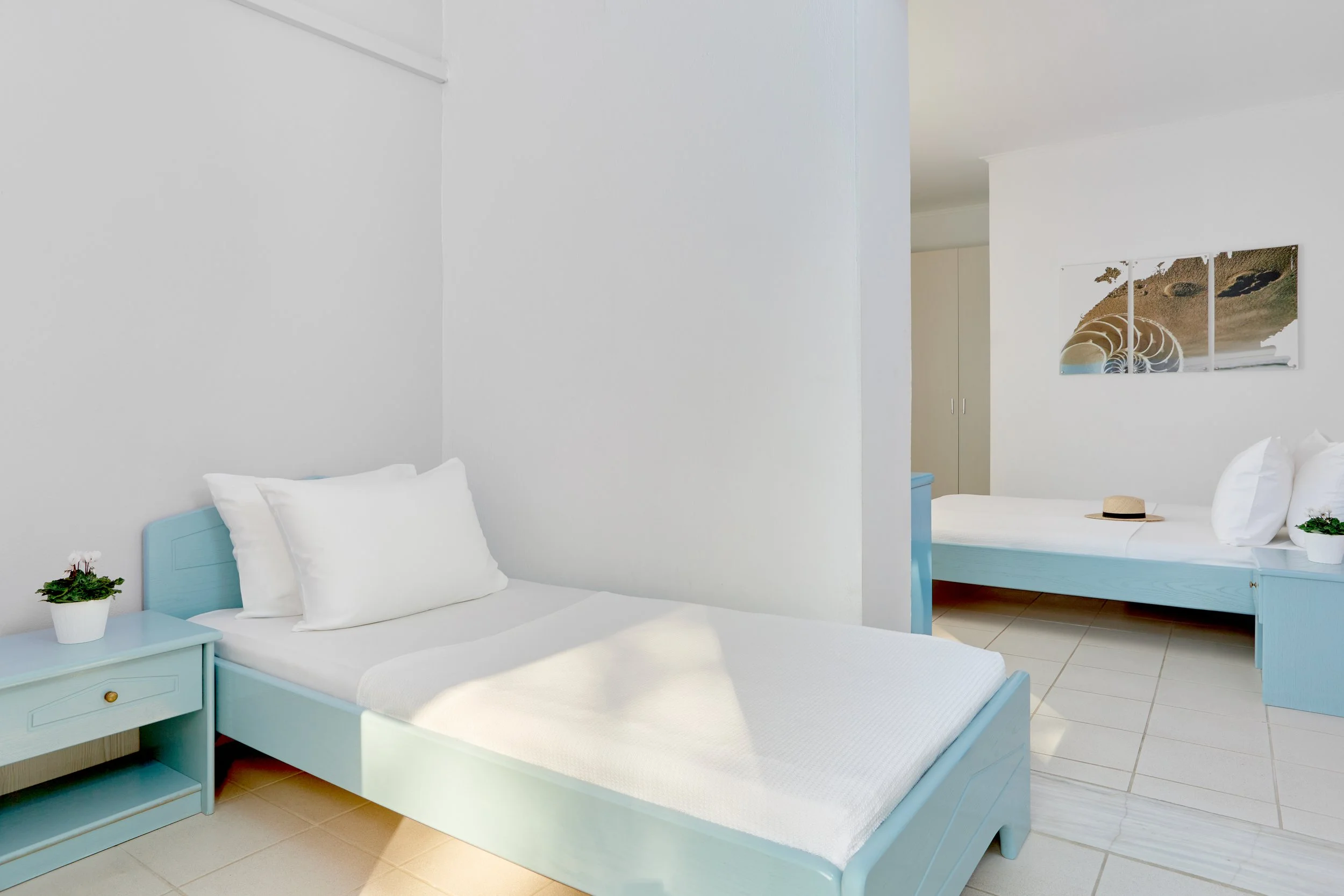 A room with white walls, two light blue beds with white bedding, one bed with a pillow and a straw hat, and a nightstand with a small potted plant. There is a large photo on the wall showing an artistic spiral and beach landscape.