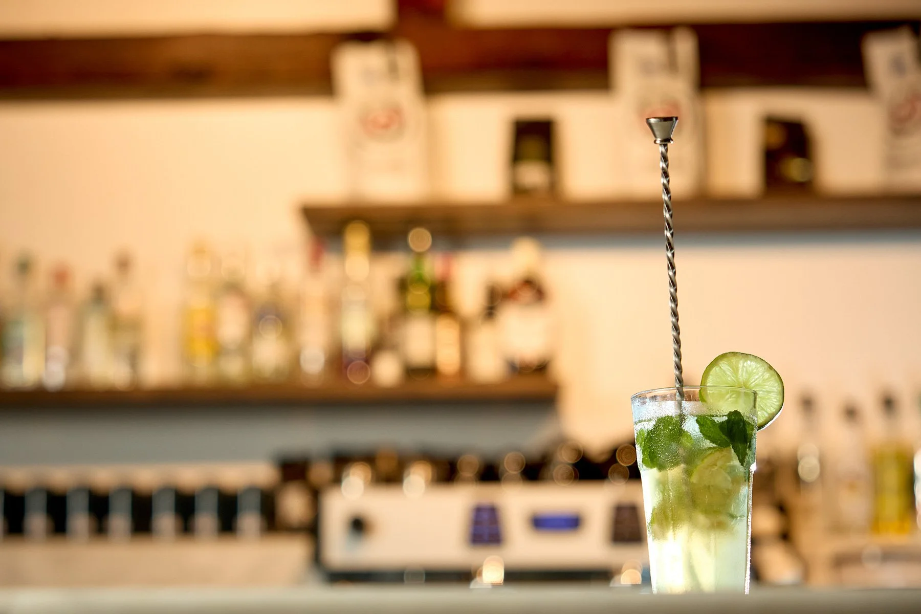 A mojito cocktail garnished with lime and mint in a tall glass, with a twisted metal stirring stick, set against a blurred bar background.