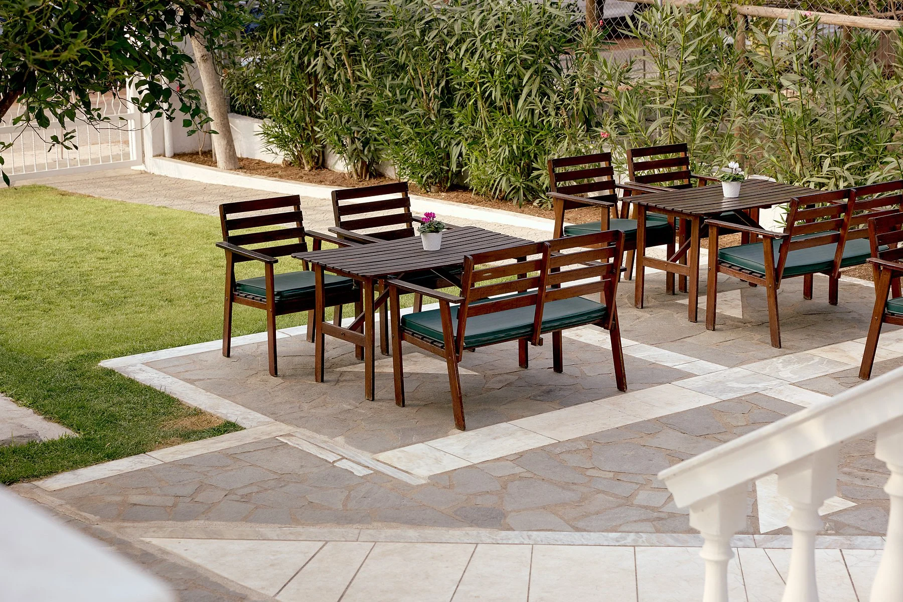 Outdoor patio with wooden tables and chairs, each table has a small potted flower, surrounded by green plants and a white fence.