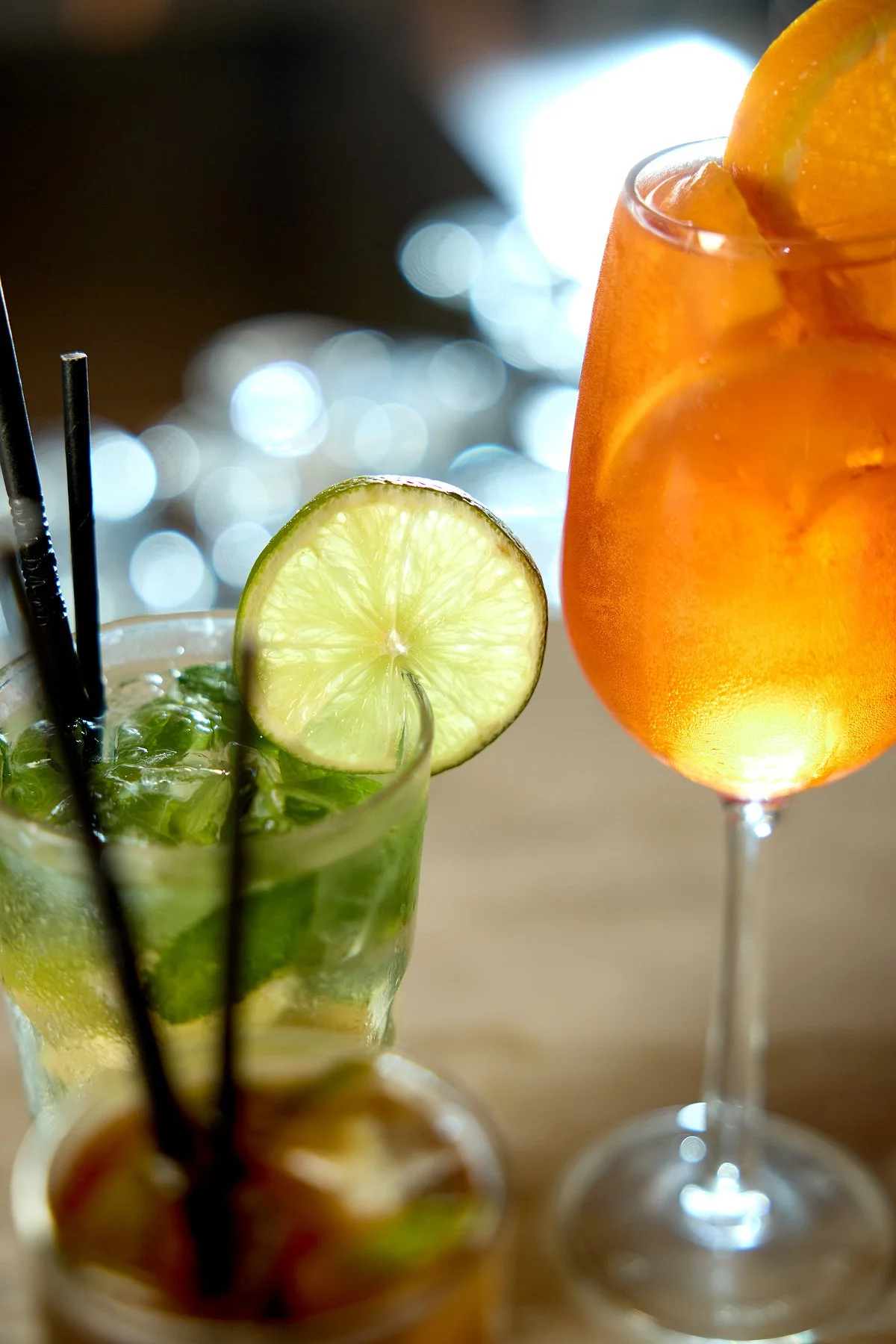 Two cocktails, one with lime and mint, and the other with oranges, on a wooden surface with blurred lights in the background.