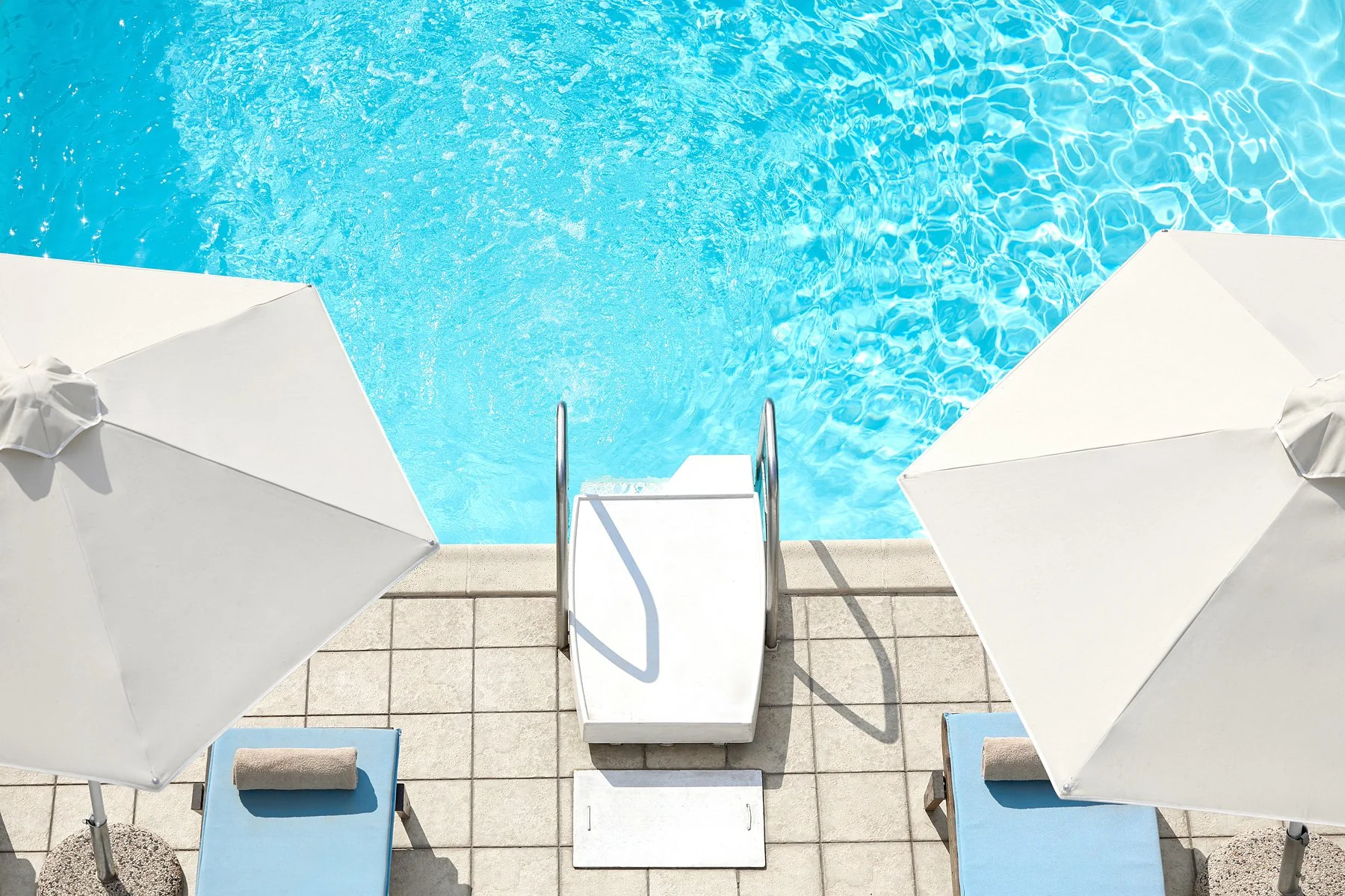 View of a swimming pool with light blue water, viewed from above. There is a small set of metal stairs leading into the pool, with two white umbrellas and two lounge chairs with blue cushions on the tiled poolside.