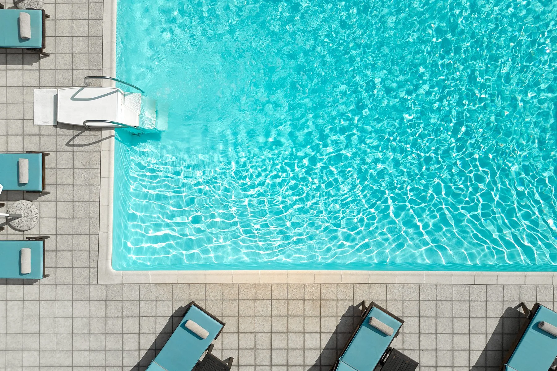 Top-down view of a swimming pool with bright blue water, surrounded by tiled decking area with several blue lounge chairs and towels.