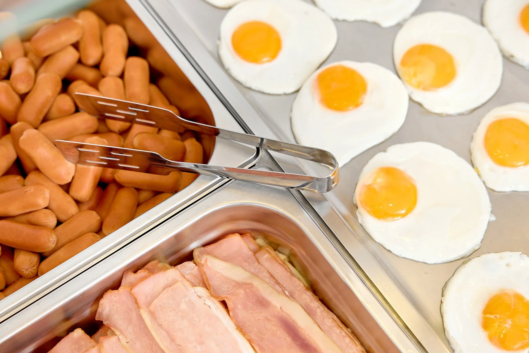 Fried eggs with runny yolks on a metal tray, cooked bacon strips, sausage links in a tray, and tongs resting on a tray of sausages.