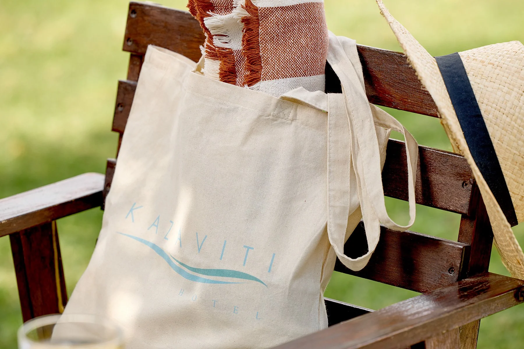 A beige tote bag hanging on the backrest of a wooden park bench, containing orange and white towels. A straw hat is on the right side of the bench.