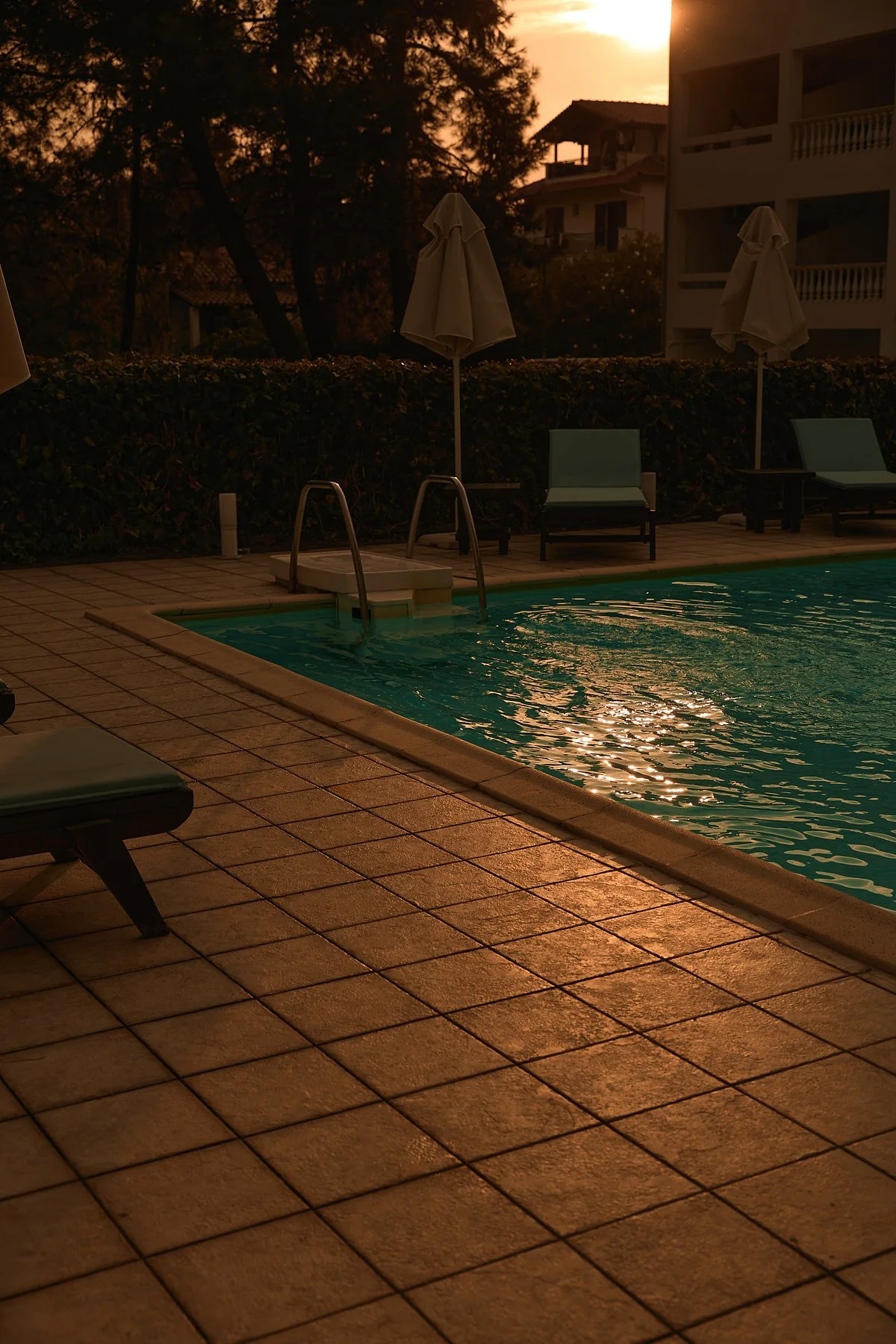 Swimming pool at sunset with pool chairs and umbrellas around, buildings and trees in the background.