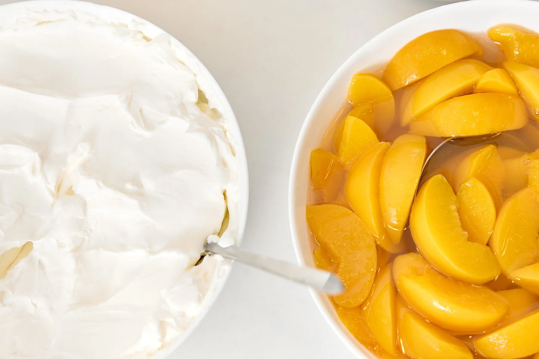 Whipped cream next to a bowl of peach slices in syrup with a spoon.