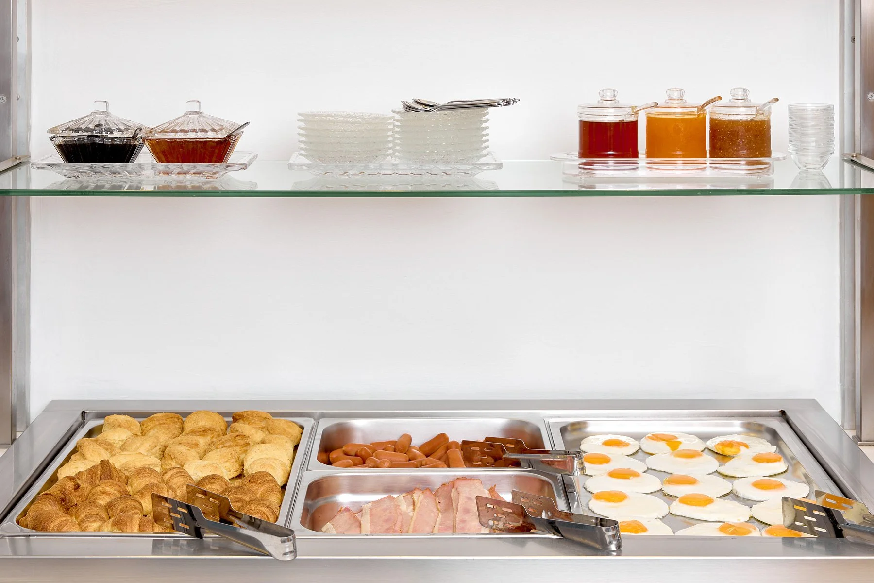 Breakfast buffet with croissants, sausage links, bacon, and fried eggs in a stainless steel serving tray, with syrup, jam, and condiments on a glass shelf above.