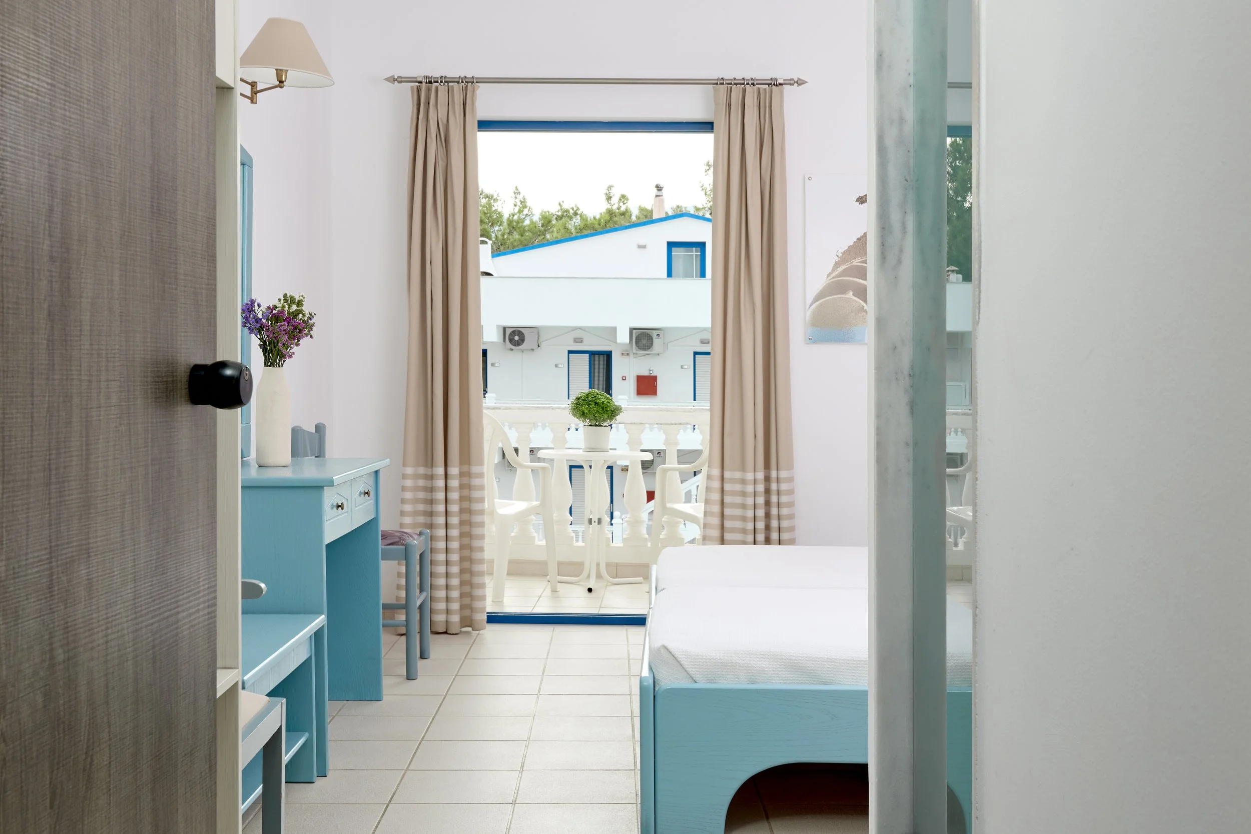 View of a seaside hotel room with a balcony, white furniture, beige curtains, and a blue and white color theme.