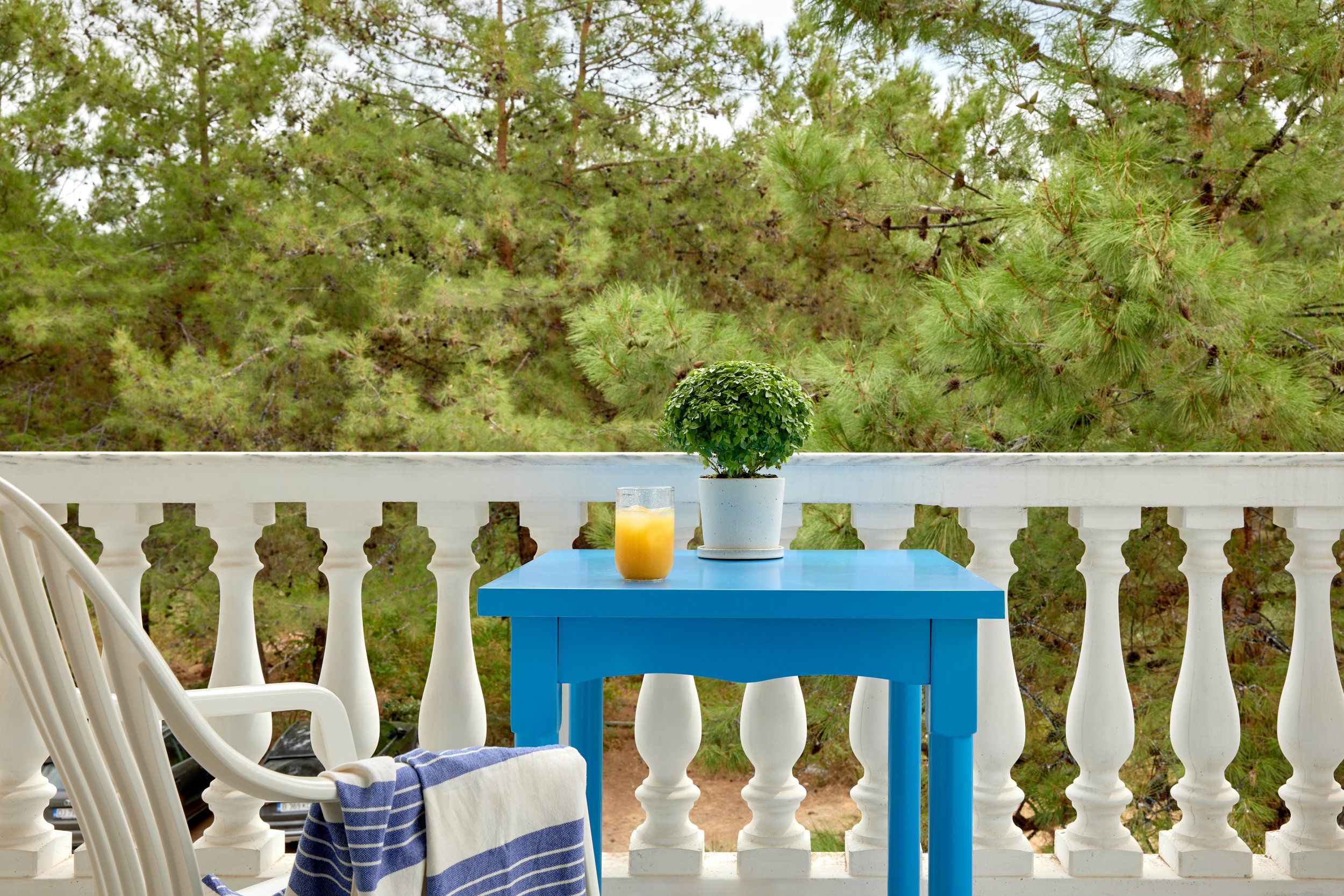 A balcony with a white railing overlooking green pine trees, a blue table with a potted plant and a glass of orange juice, and a white chair with a blue and white striped towel.