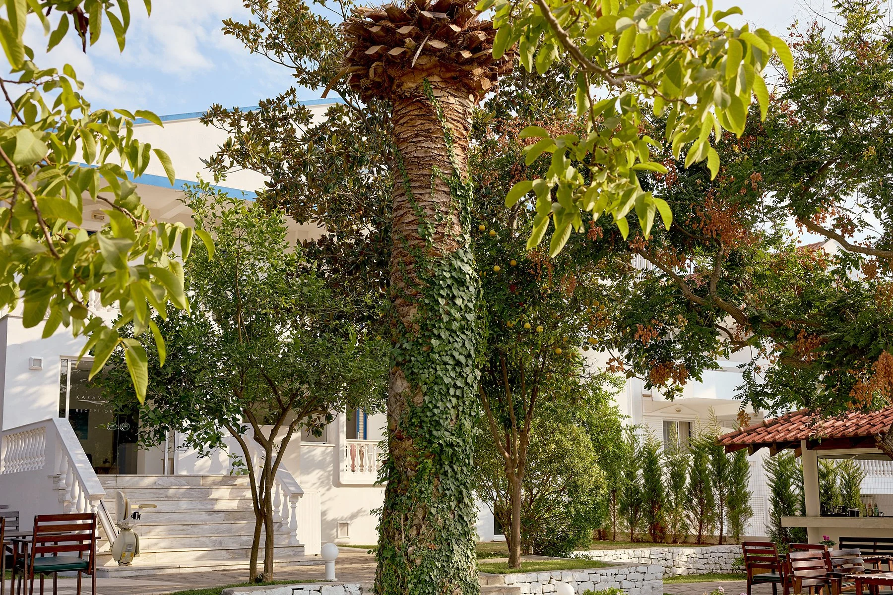 A courtyard with trees, benches, a small tiled pavilion, and a white building with stairs and balconies in the background.