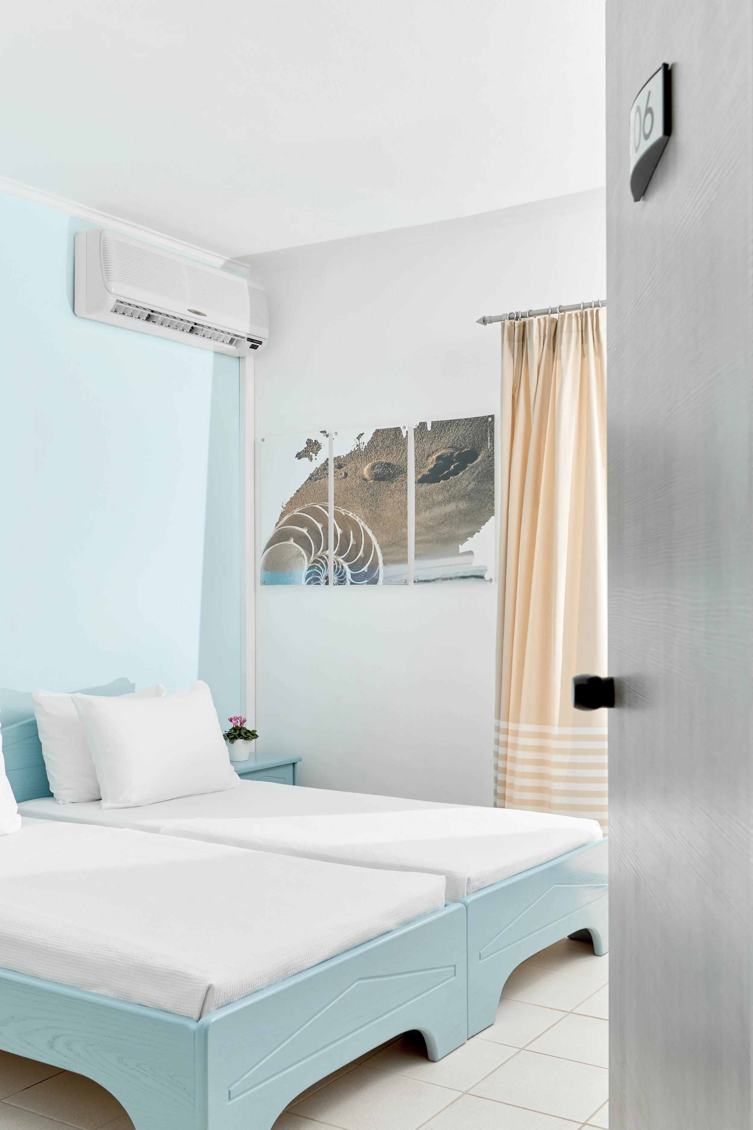 View of a minimalist hotel room with a light blue bed, white pillows, a small side table with a flowerpot, beach art on the wall, beige curtains, and an air conditioning unit on the wall.