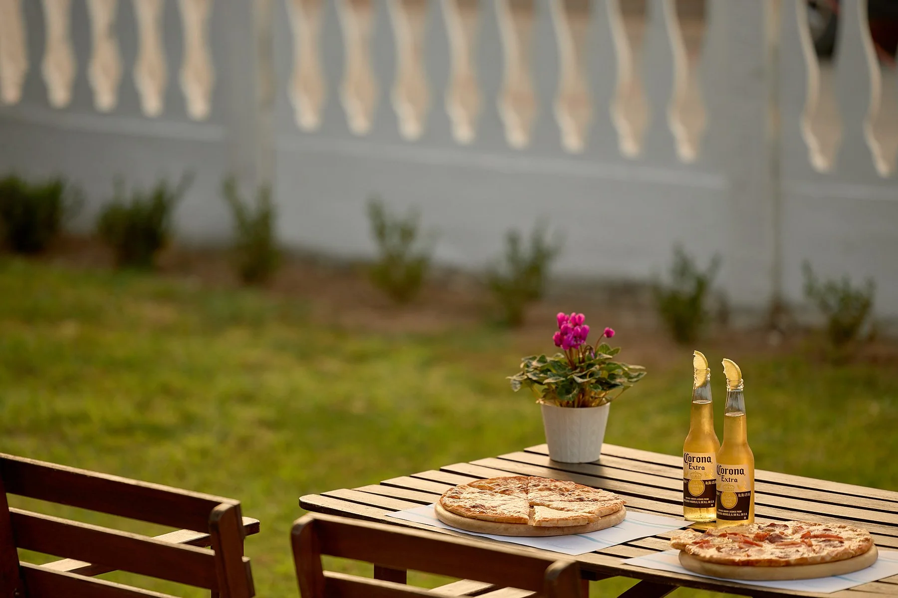 An outdoor patio table with two pizzas, two bottles of Corona Extra beer with lime wedges, and a potted pink flowering plant, with wooden chairs and a garden with grass and bushes in the background.