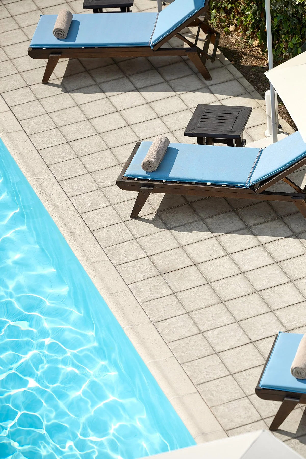 Poolside area with two lounge chairs with blue cushions, each with a rolled towel on top, a small table between the chairs, and a white umbrella. The pool water is visible on the left side.