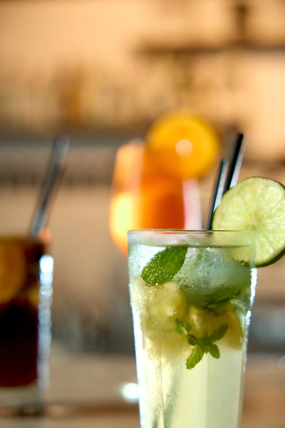 A glass of lime mojito with mint leaves and ice, garnished with a lime slice, in focus with two blurred cocktails in the background.