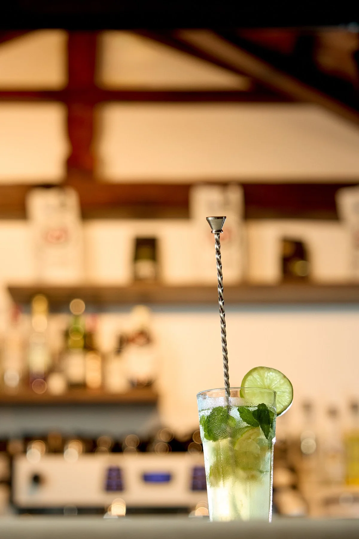 A cocktail glass filled with a lime and mint drink, garnished with a lime slice on the rim, and a curly straw, set against a blurred bar background.