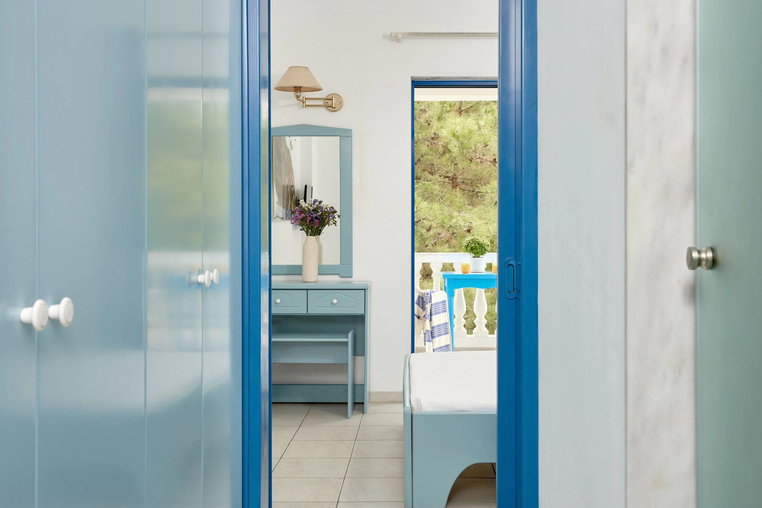 View through the doors of a white and blue bedroom with a bed, dresser, mirror, flowers, and balcony with trees outside.