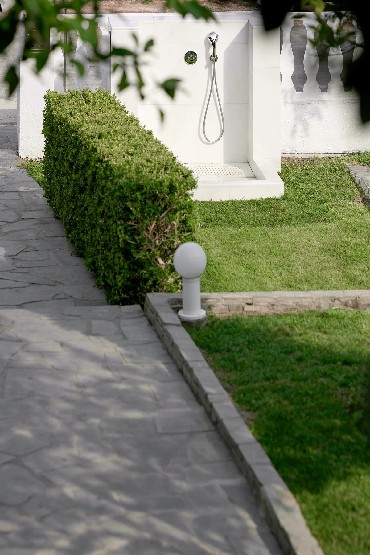 Outdoor shower area with white walls, a silver shower head and hose, and a grassy lawn, bordered by a stone pathway and a green hedge.