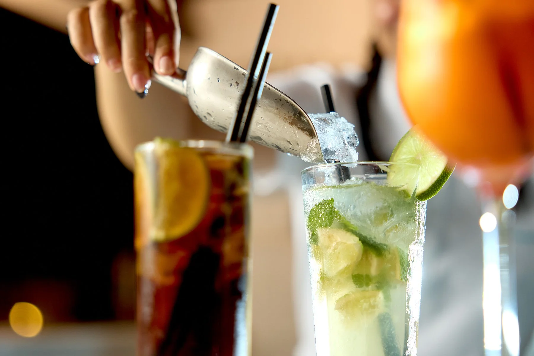A bartender is pouring a clear drink into a glass filled with lime slices and ice, with a slice of lime on the rim. In the foreground, there is a partially visible drink with a lemon wedge, and in the background, another cocktail with an orange slice