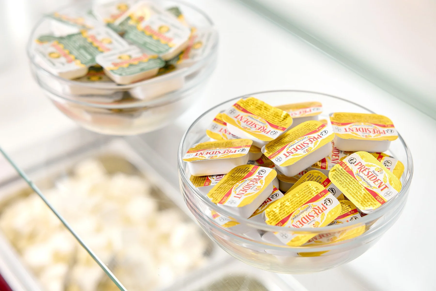 Two glass bowls, one in the foreground filled with individual servings of President brand margarine, and a second bowl in the background filled with many more margarine containers, on a glass counter.