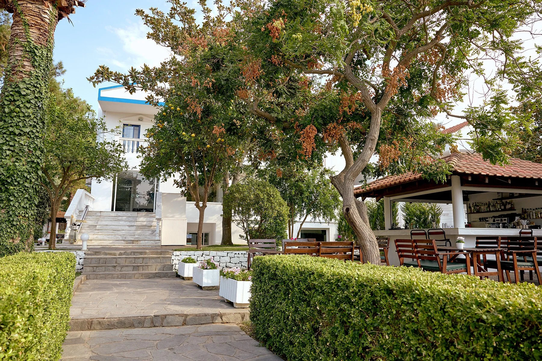 Outdoor garden area with trees, shrubs, potted plants, a stone pathway, a set of stairs leading to a white house, and a shaded seating area with wooden tables and chairs.