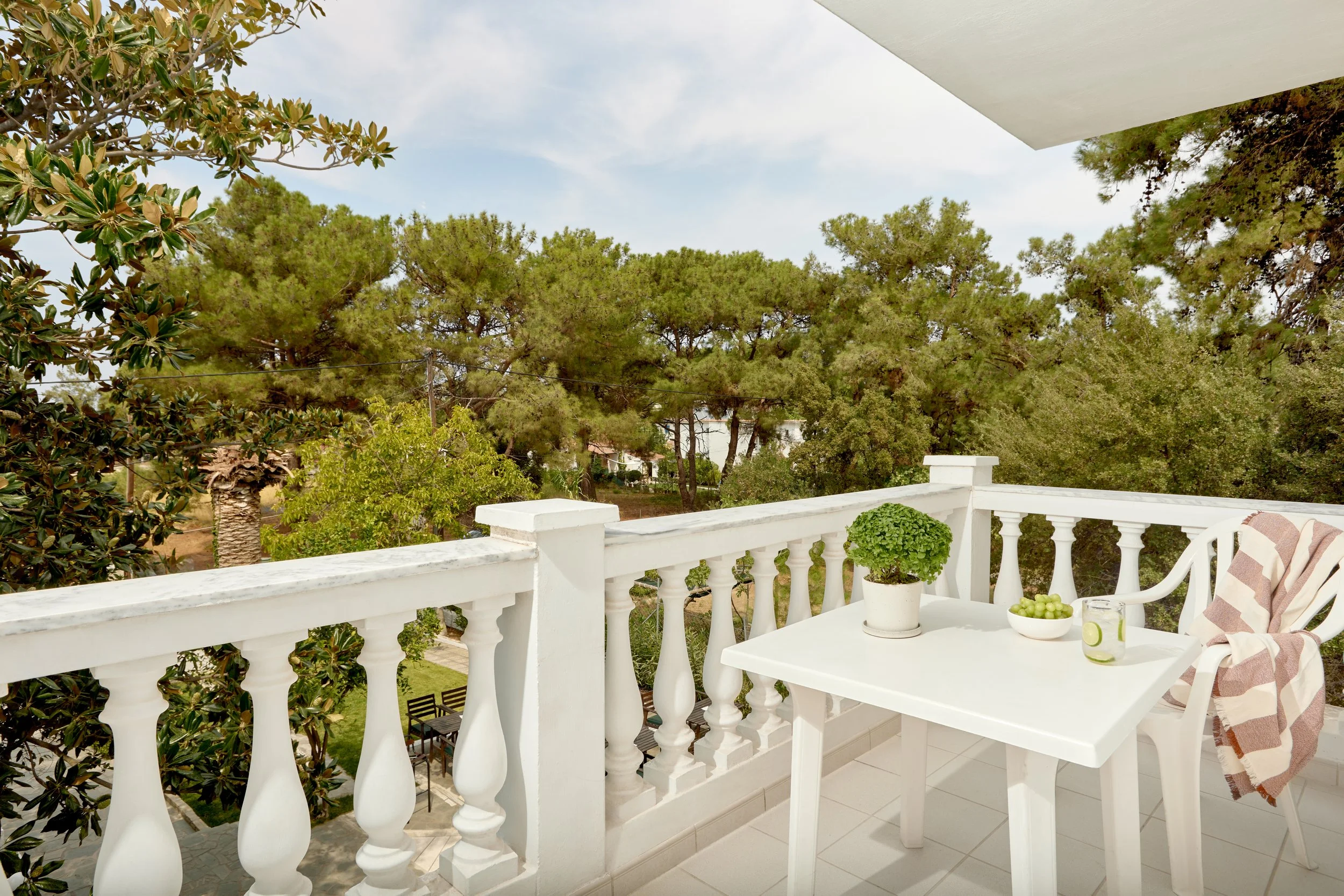 Balcony with white railing overlooking trees, with a small white table holding a potted plant, a bowl of green grapes, and a glass of water. There is a chair with a striped cloth draped over it.
