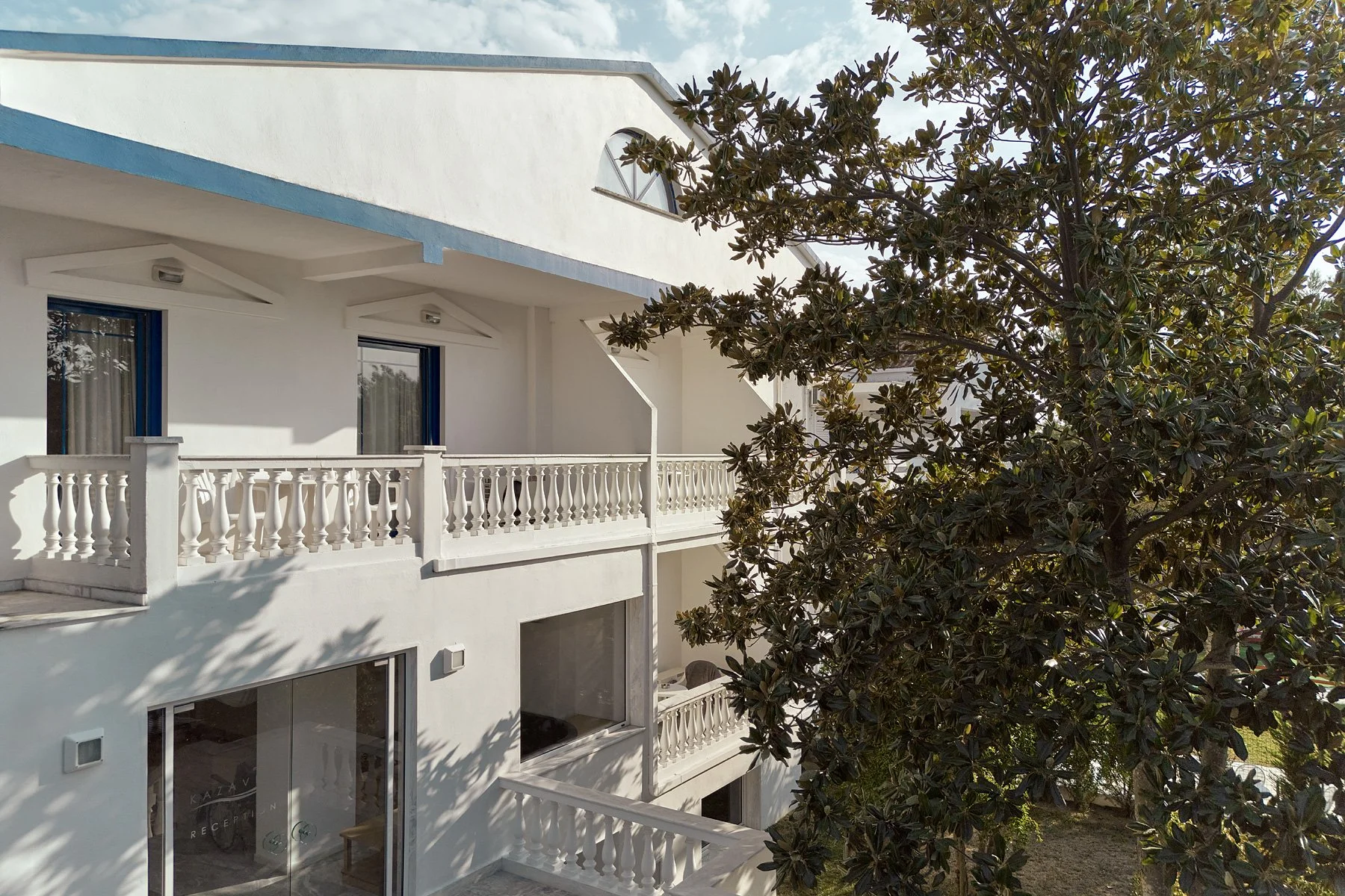 White multi-story residential building with balconies, blue window frames, and a large green tree in front.