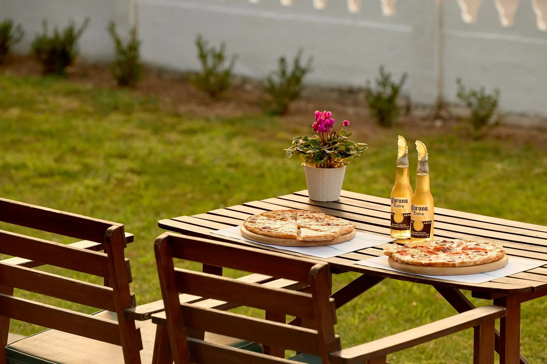 Outdoor patio table with two pizzas on white paper sheets, two bottles of Corona beer with lemon wedges, and a potted pink flowering plant in the background, with wooden chairs and a grassy yard.