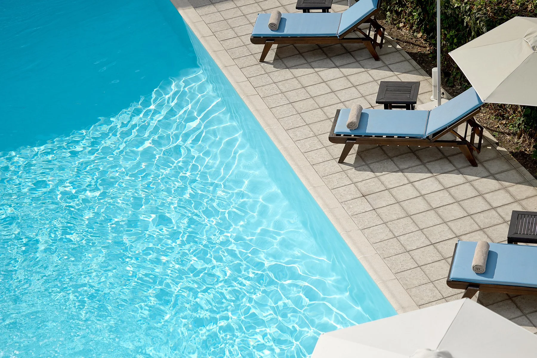 Swimming pool with clear blue water and four lounge chairs with blue cushions and rolled towels, shaded by large white umbrellas, on a tiled poolside area.