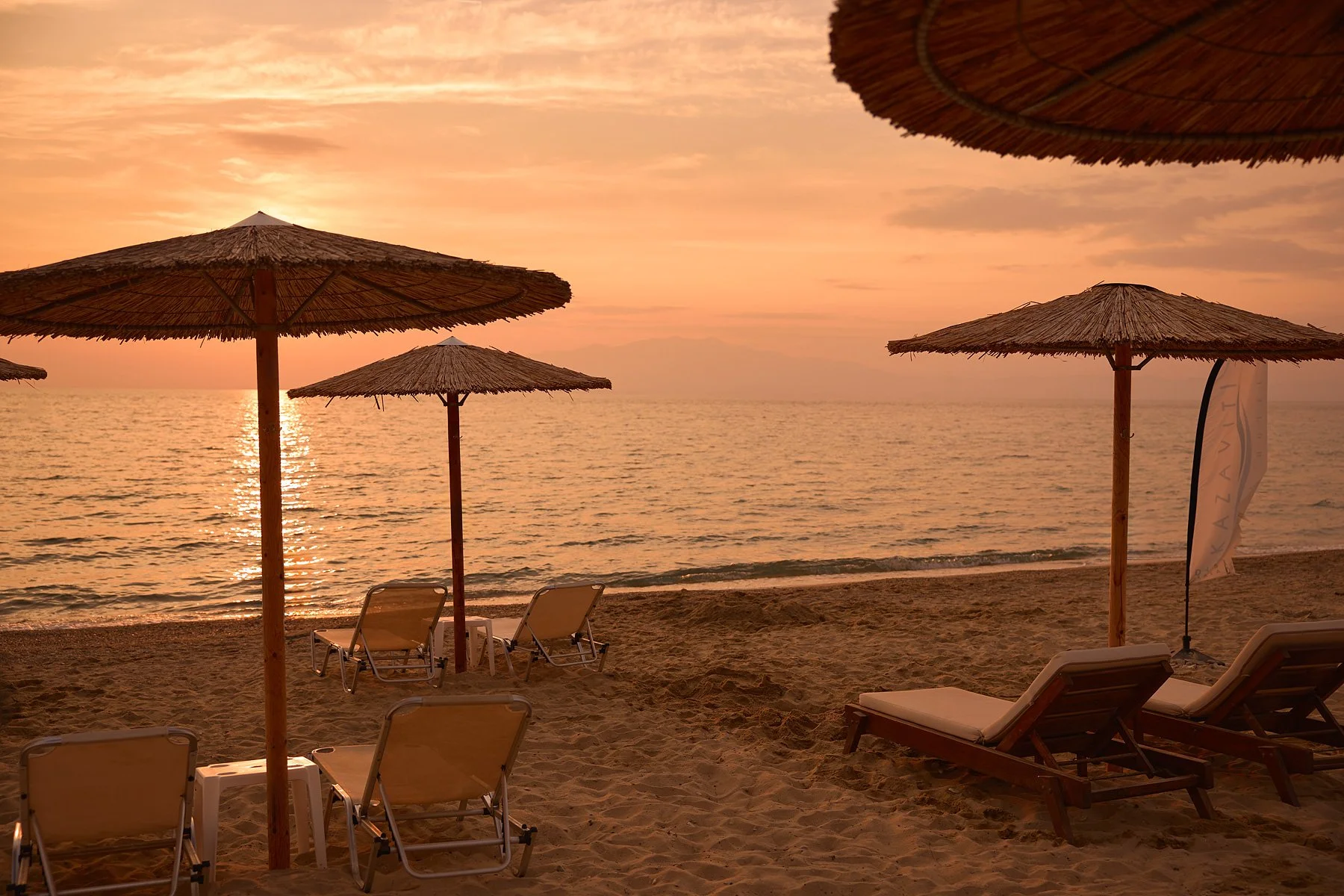Stony beach at sunset with several empty lounge chairs and thatched umbrellas.