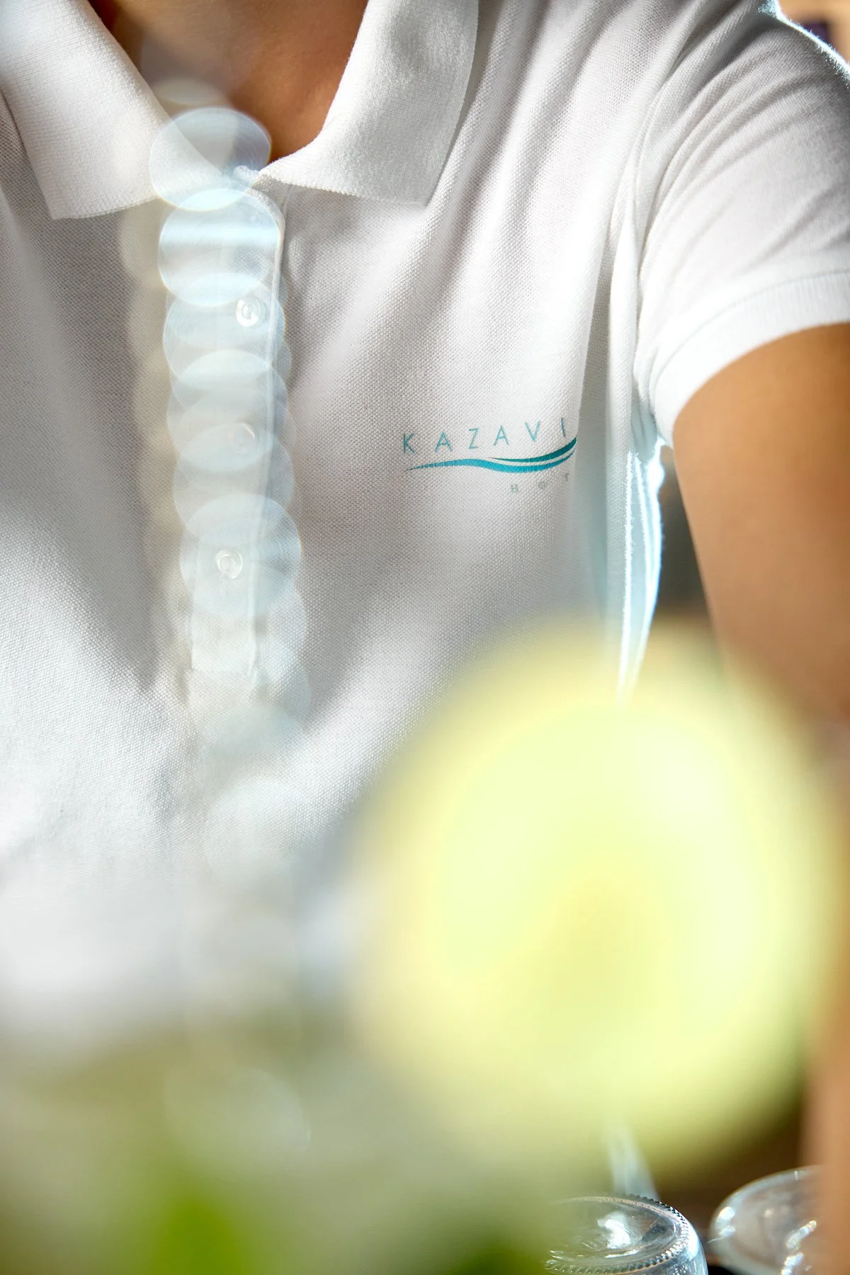 Close-up of a person wearing a white polo shirt with a logo that says 'KAZAVI' on the left side. The photo is taken at an angle with a blurred yellow object in the foreground and a soft light creating bokeh effects.