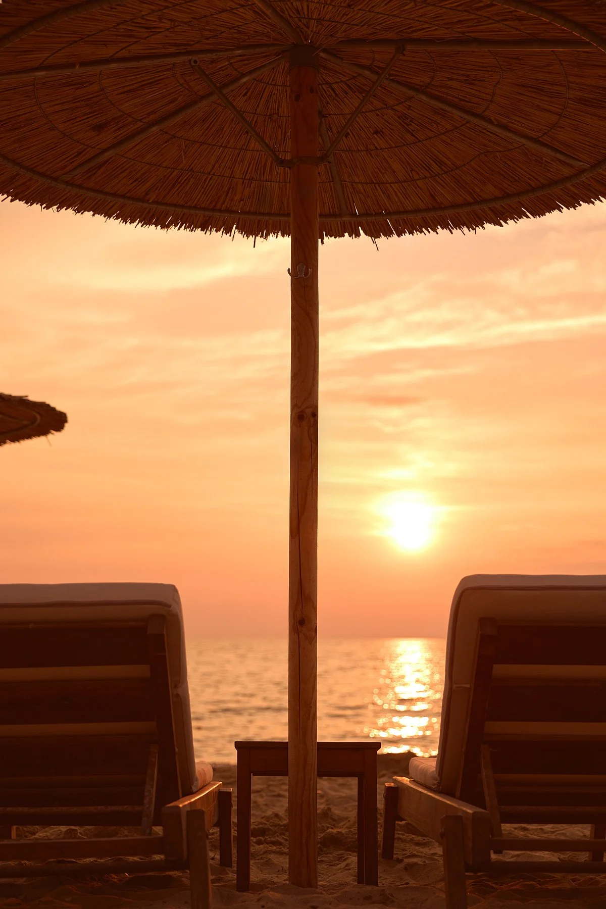 Beach scene at sunset with two lounge chairs, a small table, a large straw beach umbrella, calm ocean, and a colorful sky.