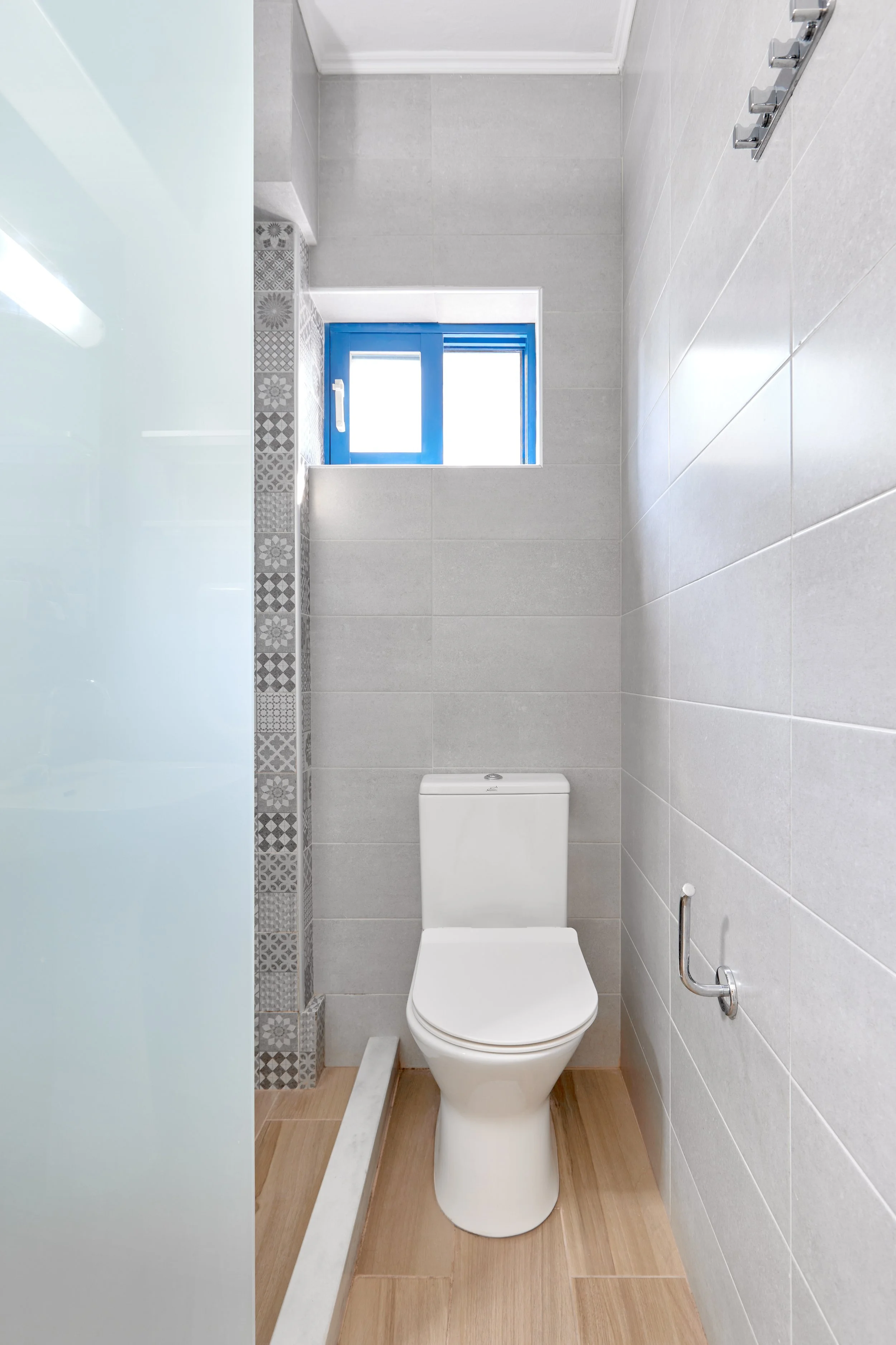 Small, modern bathroom with a white toilet, light gray tiled walls, a small window with blue trim, and a partial glass shower enclosure.