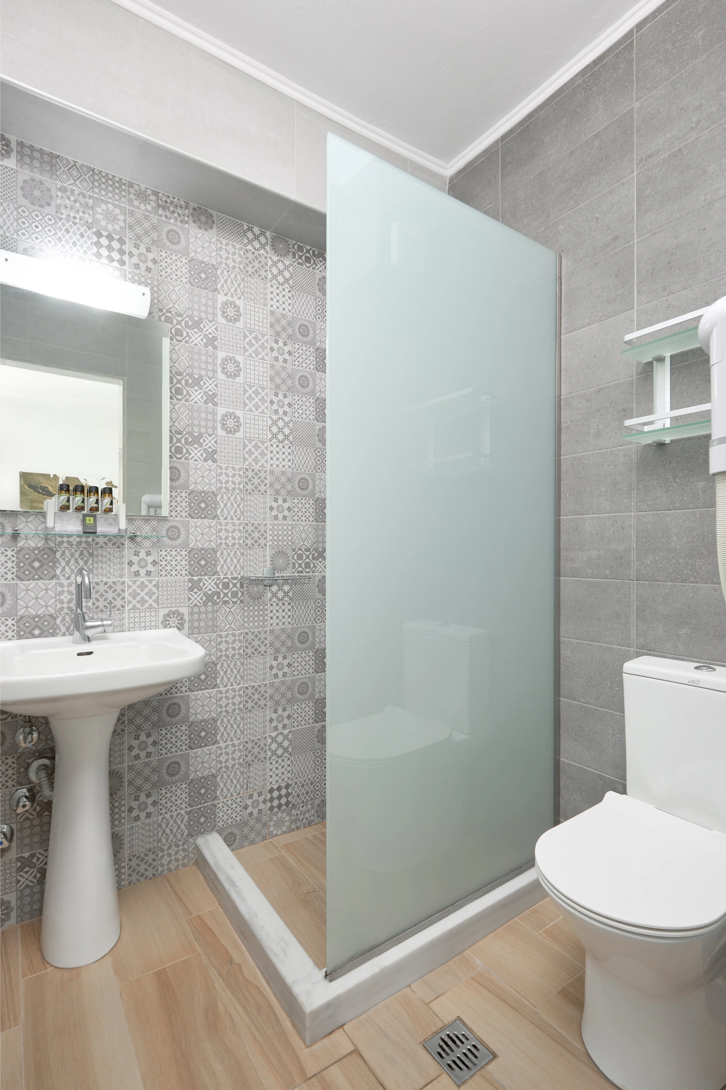 Small modern bathroom with a pedestal sink, a frosted glass shower partition, a toilet, and patterned gray tiles on the wall.