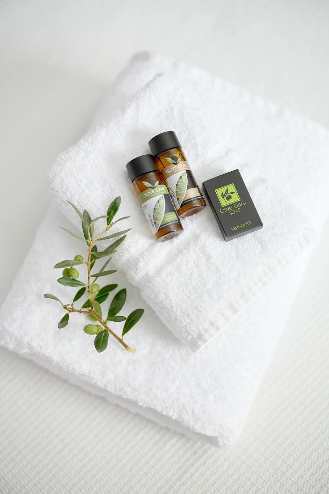 Two small brown bottles labeled olive care with black caps, a black box labeled olive care soap, and a sprig of green olives on a white rolled towel placed on another white textured towel.