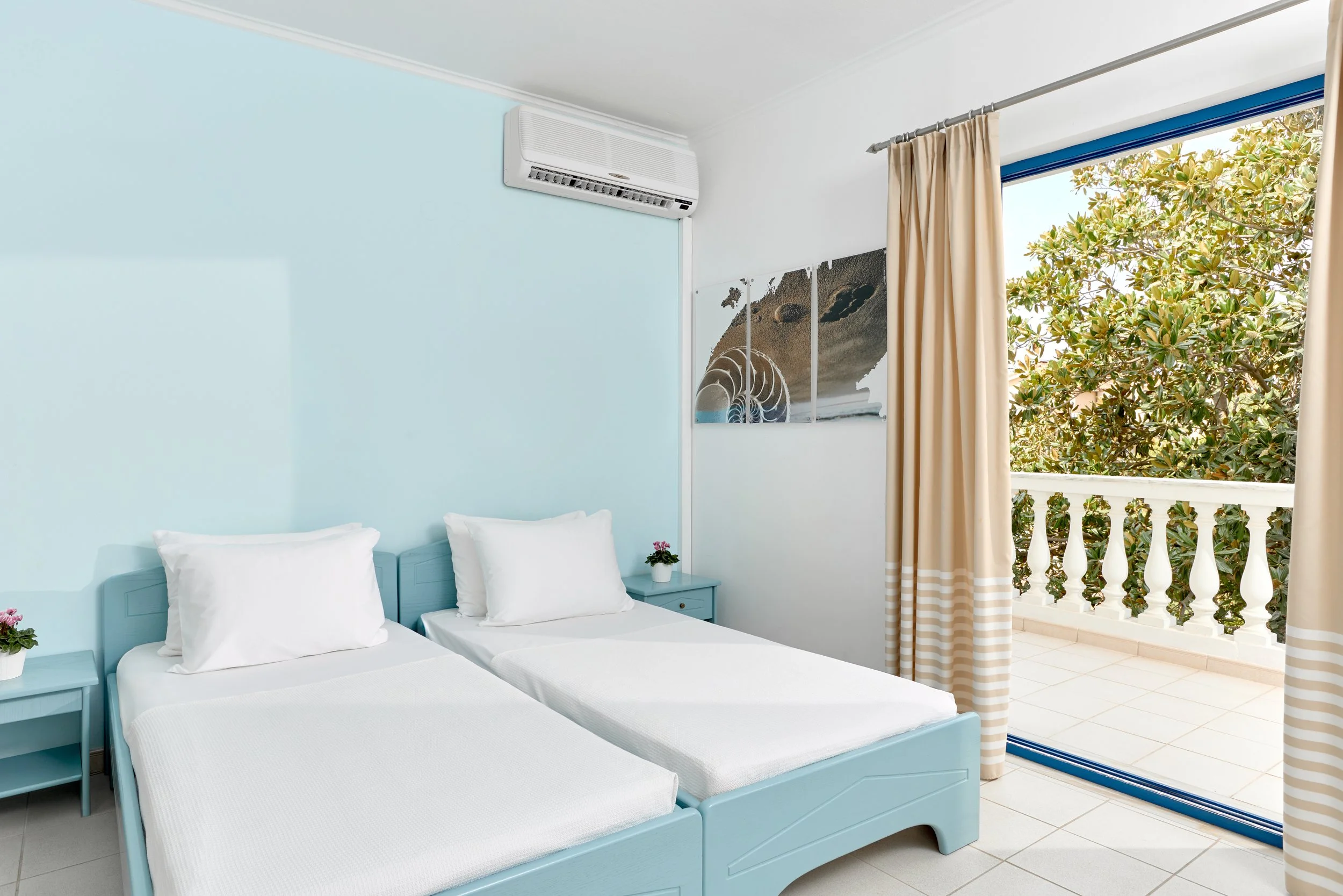 A bedroom with two single beds with white linens, a blue nightstand, tropical curtains, and a balcony with greenery and ocean view.