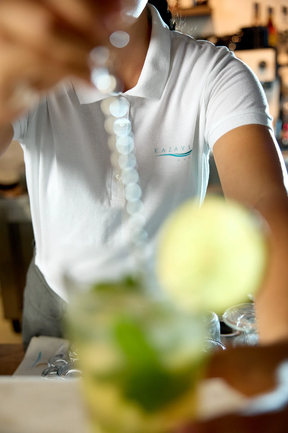 Person wearing a white polo shirt with a logo, reaching over a table with blurred green limes and a glass of some beverage in front.