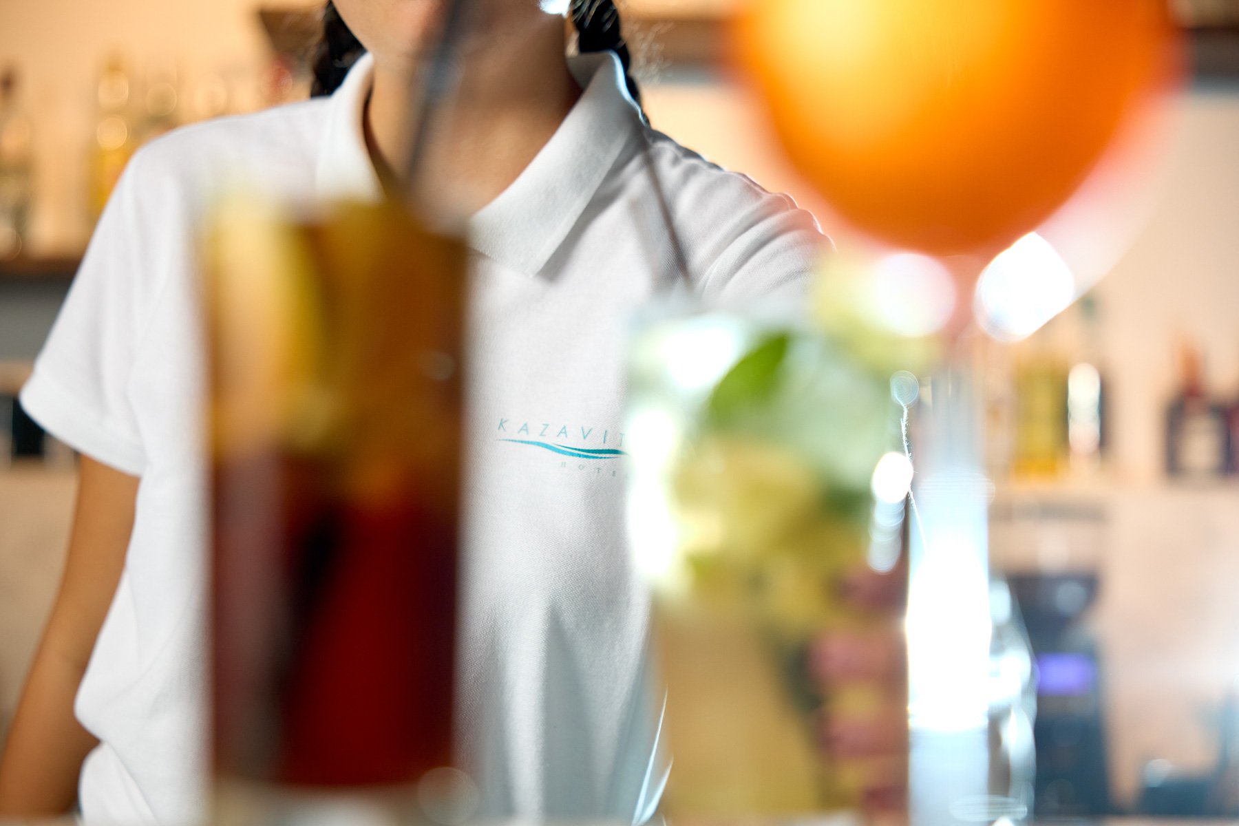 A person behind a counter with colorful drinks and oranges in the foreground, out of focus.