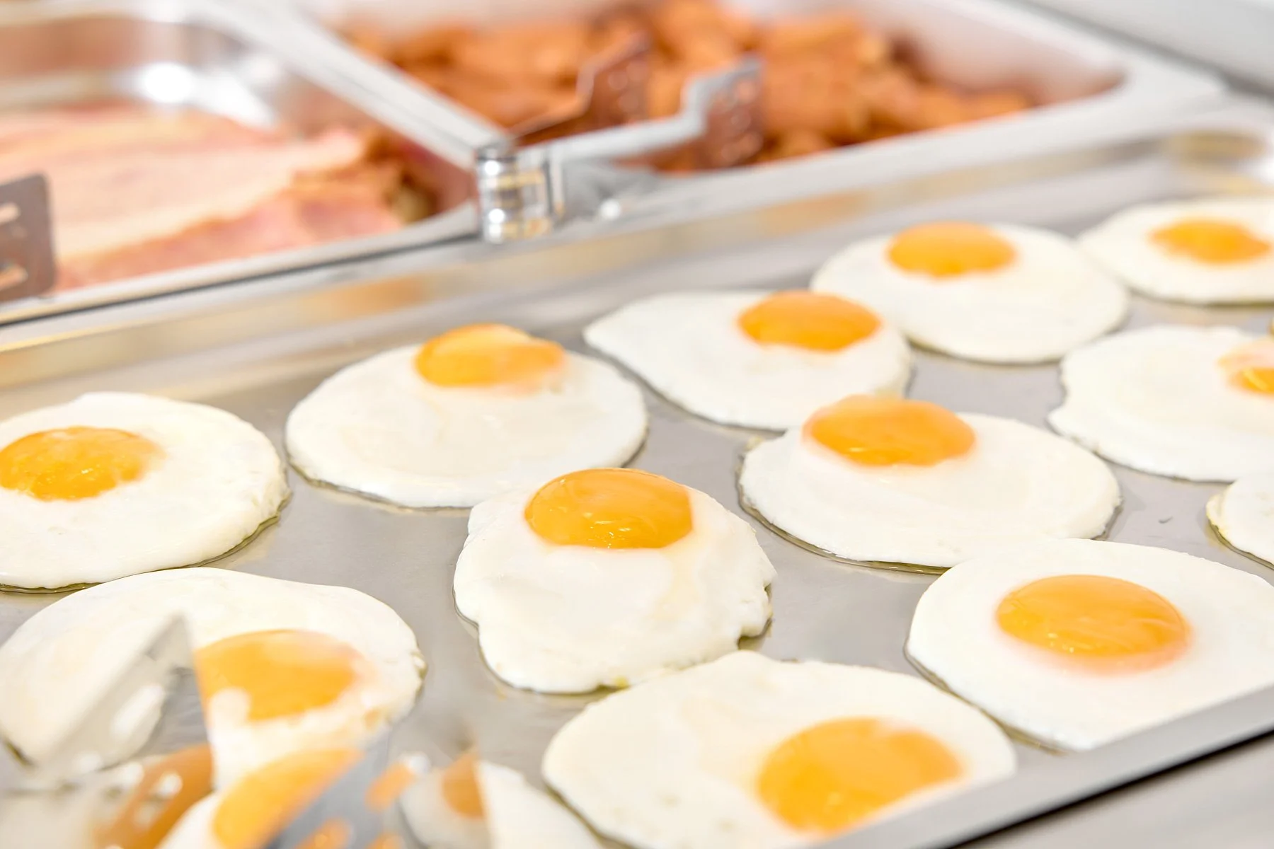 Close-up of cooked sunny-side-up eggs on a metal baking sheet with a tray of bacon and cooked breakfast meats in the background.