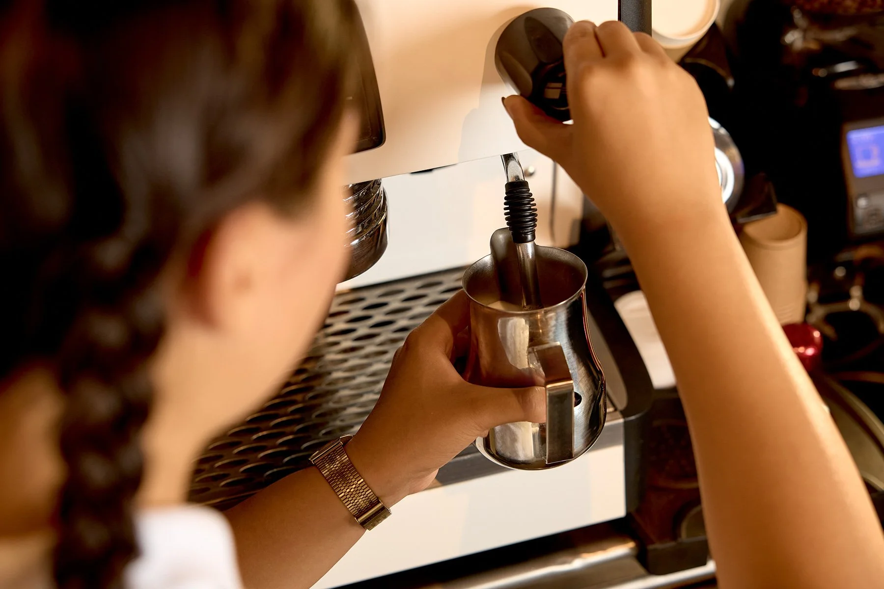 A person with a braid and wearing a bracelet is using a coffee machine to make espresso, holding a metal milk frothing pitcher under the steam wand.