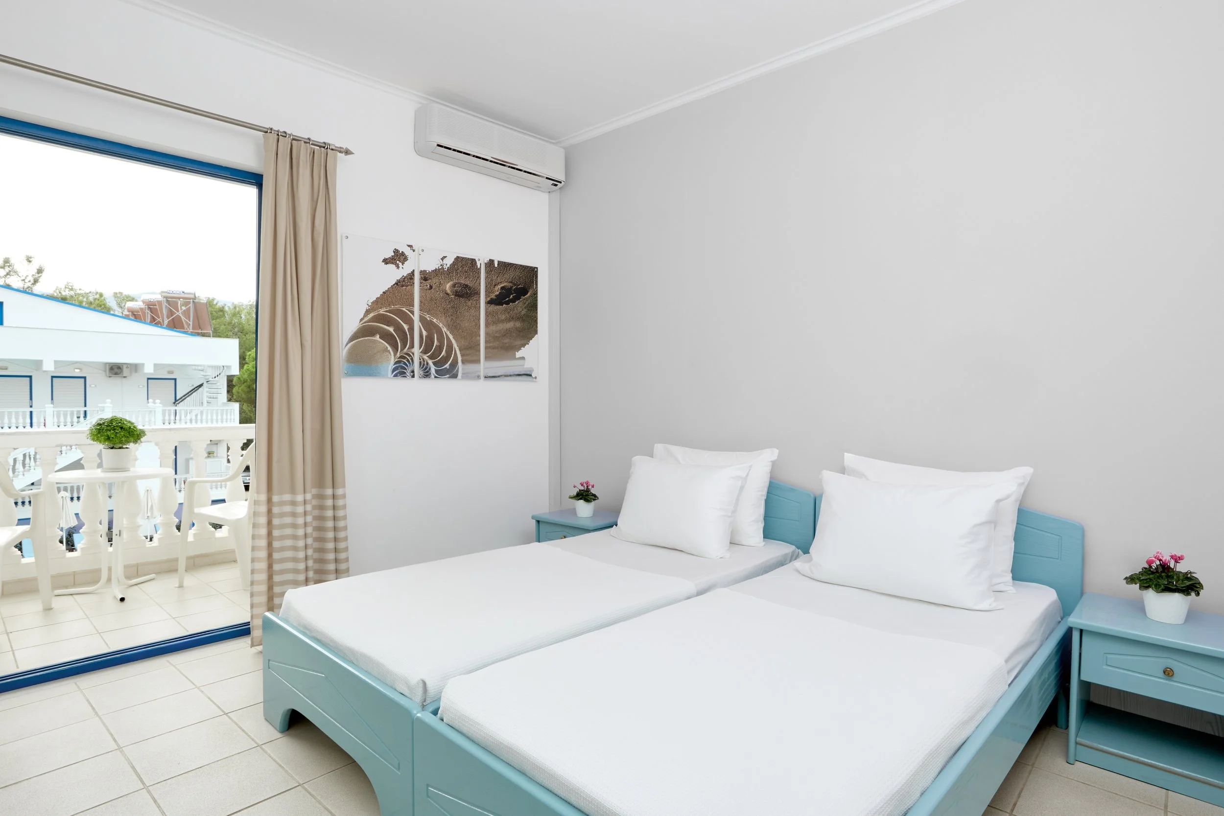 A bright hotel room with two single beds with white linens and light blue frames, small light blue nightstands with pink flowers, a large window with beige curtains, a balcony with outdoor furniture, and framed beach-themed artwork on the wall.