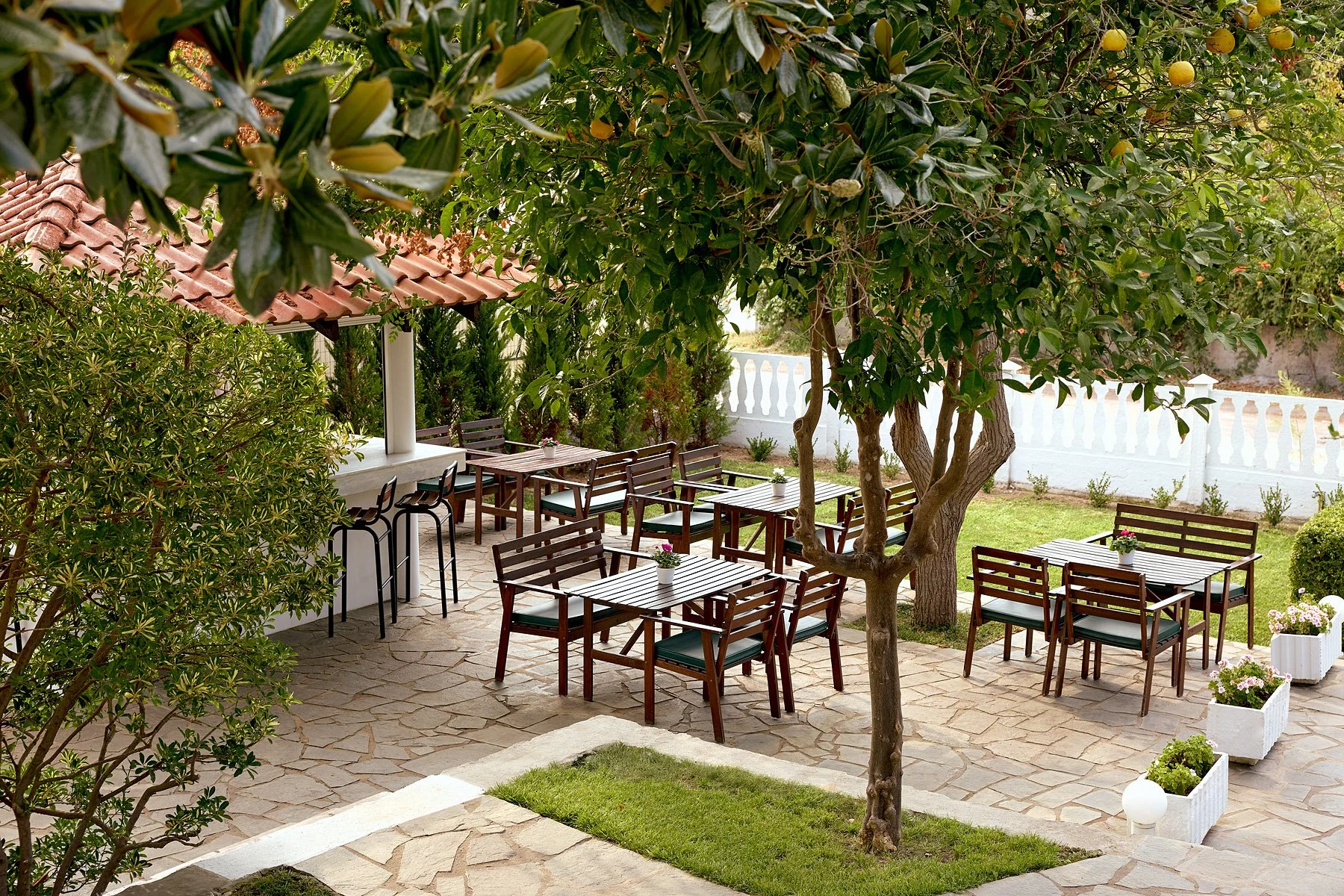 Outdoor patio area with wooden tables and chairs, white planters with pink flowers, shaded by trees, with a white fence in the background.