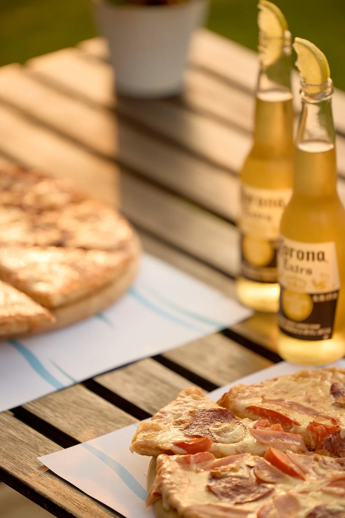 Slices of pepperoni pizza on white paper on a wooden table, with bottles of Corona beer with lemon wedges and a cup in the background.